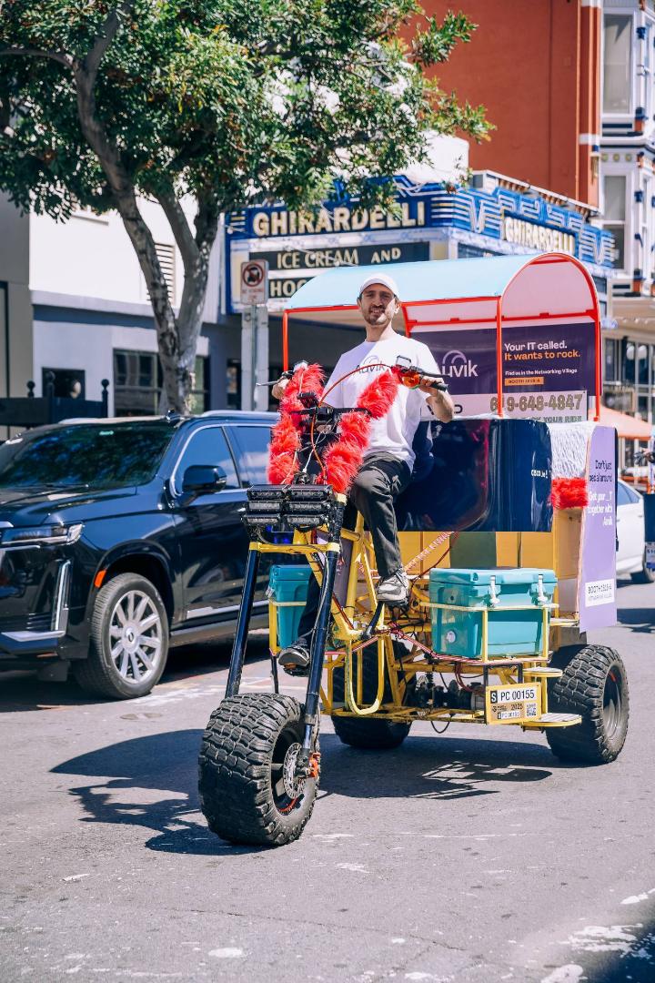 A smiling driver on an oversized, Auvik-wrapped electric chariot with monster tires and red fur trim in the Gaslamp Quarter