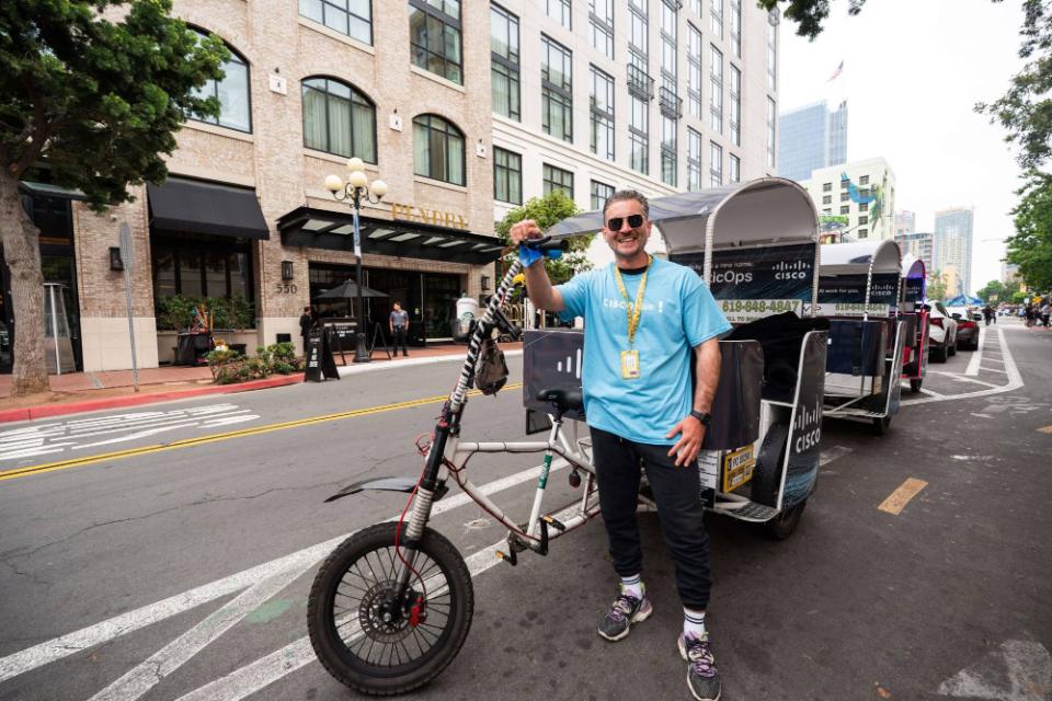 A smiling driver in a Cisco Live shirt beside a line of wrapped pedicabs outside the Pendry hotel