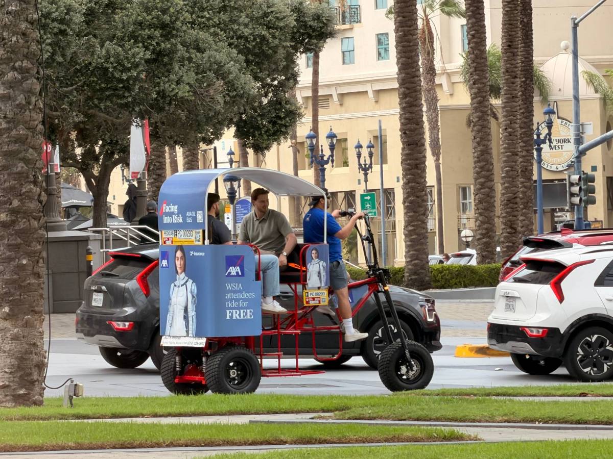 Two riders in a Power Chariot wrapped with a sponsor ad reading WSIA attendees ride for free, driving past the Convention Center area
