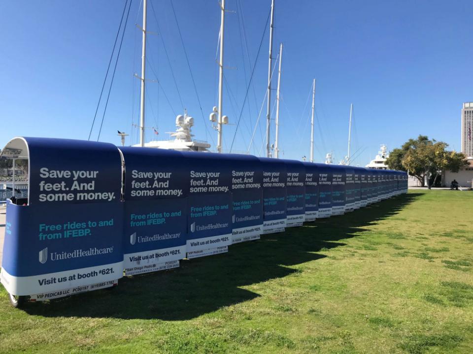 A long line of pedicabs wrapped for UnitedHealthcare parked on the lawn by the waterfront marina